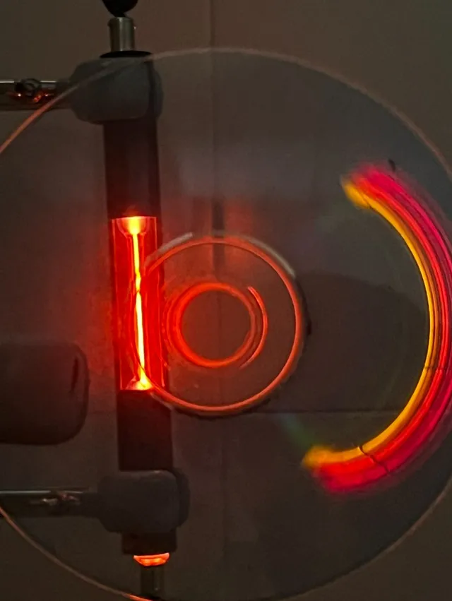Hecho en clase: un tubo de descarga de neón reflejado en un CD. El surco del disco separa la luz y asoman sus rayas rojas y naranjas.