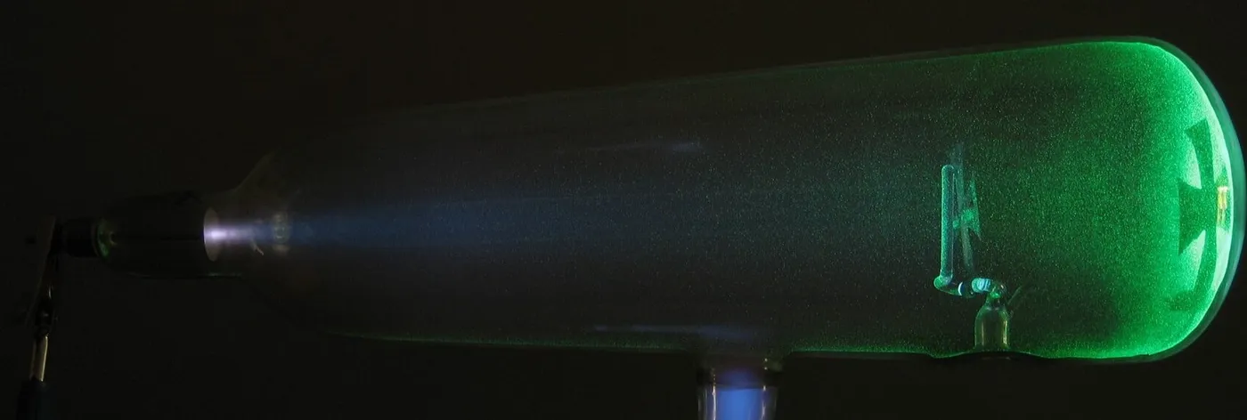 Un tubo de rayos catódicos encendido: el haz sale del electrodo negativo, a la izquierda, hace brillar el vidrio de verde y la cruz metálica deja su sombra al fondo
