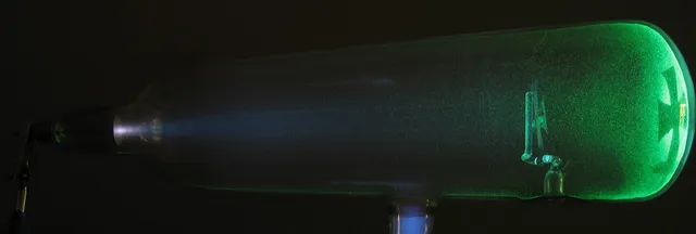 Un tubo de rayos catódicos encendido: el haz sale del electrodo negativo, a la izquierda, hace brillar el vidrio de verde y la cruz metálica deja su sombra al fondo